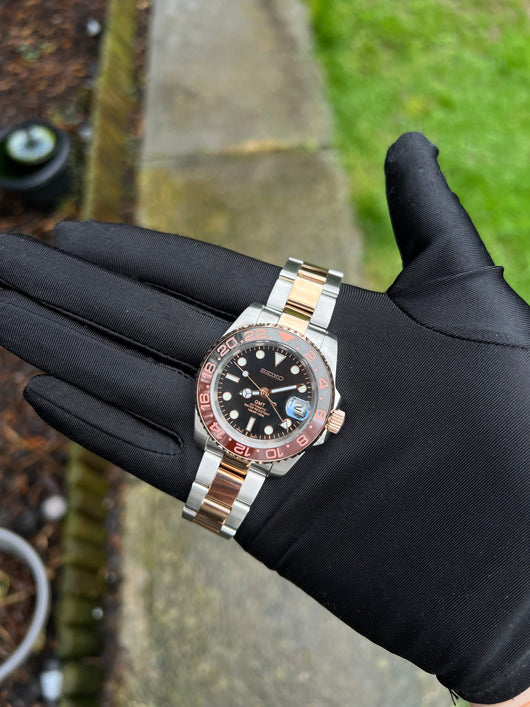 GMT (Rootbeer) Seiko NH34 GMT automatic mod watch. A full watch with a black dial and a black and rose gold bezel on a two tone oyster bracelet held in a glove.