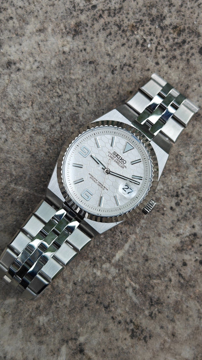 Land-Dweller Seiko NH35 automatic mod watch. A top down view of a white dial watch with a silver fluted bezel and metal link bracelet on stone.