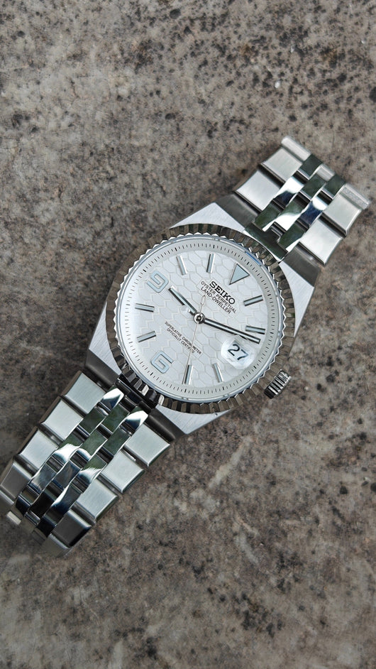 Land-Dweller Seiko NH35 automatic mod watch. A top down view of a white dial watch with a silver fluted bezel and metal link bracelet on stone.