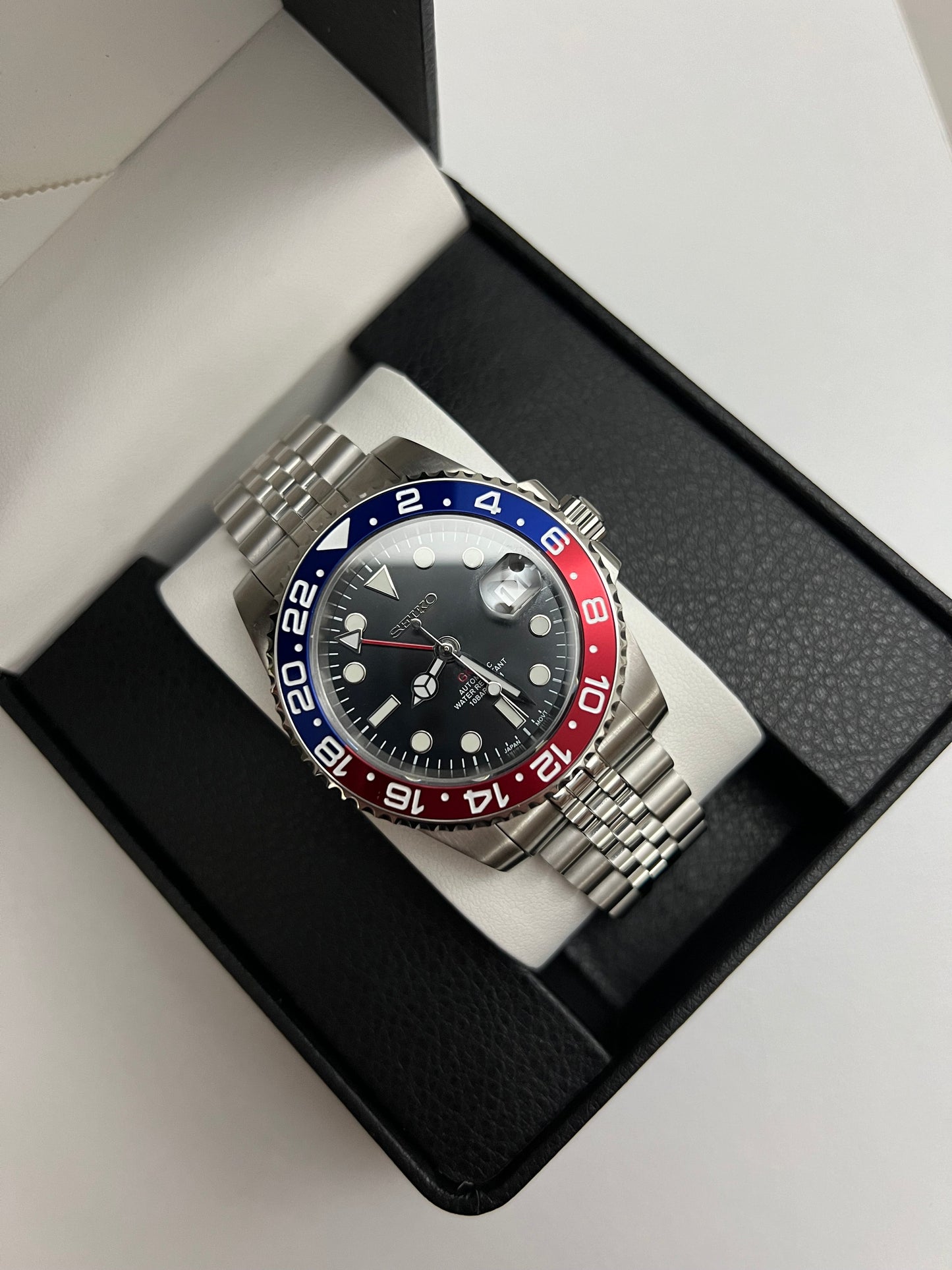 GMT (Pepsi) Seiko NH34 GMT automatic mod watch. A full watch shot inside a box shows a black dial with a blue and red bezel on a silver jubilee bracelet.
