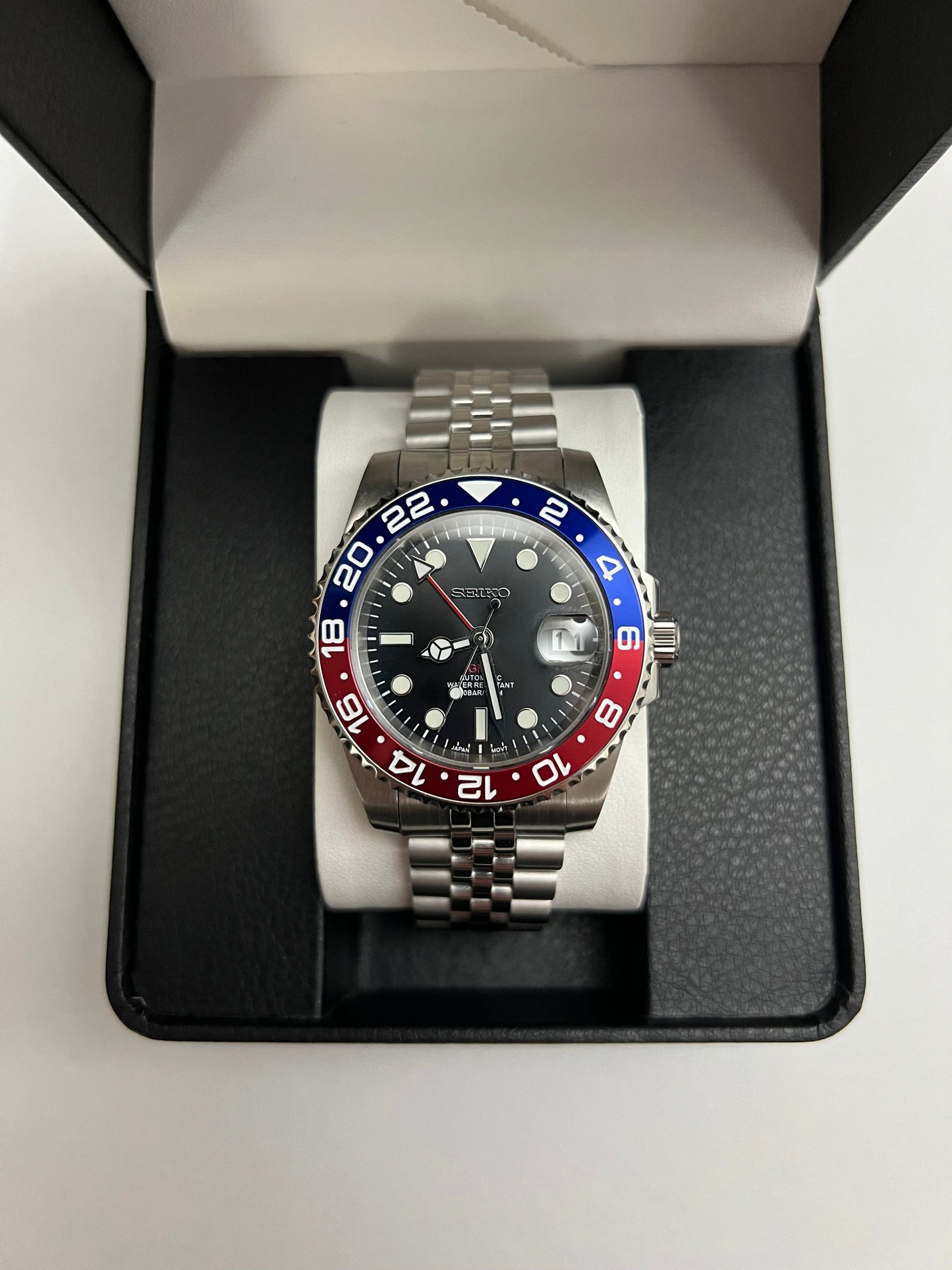 GMT (Pepsi) Seiko NH34 GMT automatic mod watch. A full watch shot inside a box shows a black dial with a blue and red bezel on a silver jubilee bracelet.