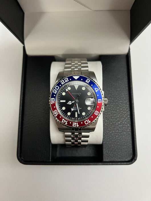 GMT (Pepsi) Seiko NH34 GMT automatic mod watch. A full watch shot inside a box shows a black dial with a blue and red bezel on a silver jubilee bracelet.