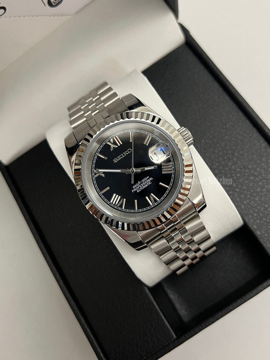 Datejust (Black Roman) Seiko NH35 automatic mod watch. A black dial watch with a silver fluted bezel and jubilee bracelet inside a box in a high angle shot.