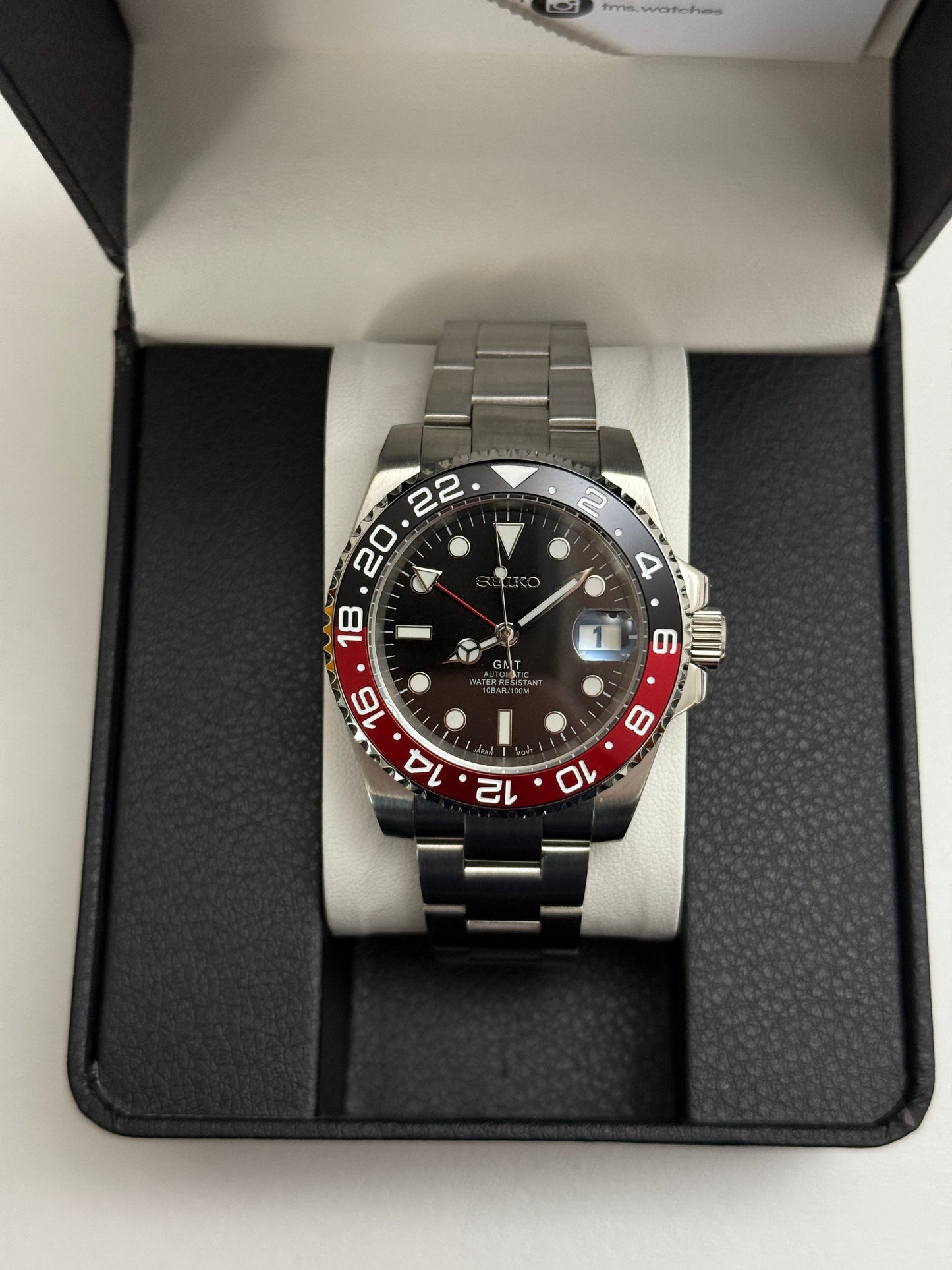 GMT (Coke) Seiko NH34 GMT automatic mod watch. A black dial watch with a black and red bezel on a metal link bracelet inside a box in a top down shot.