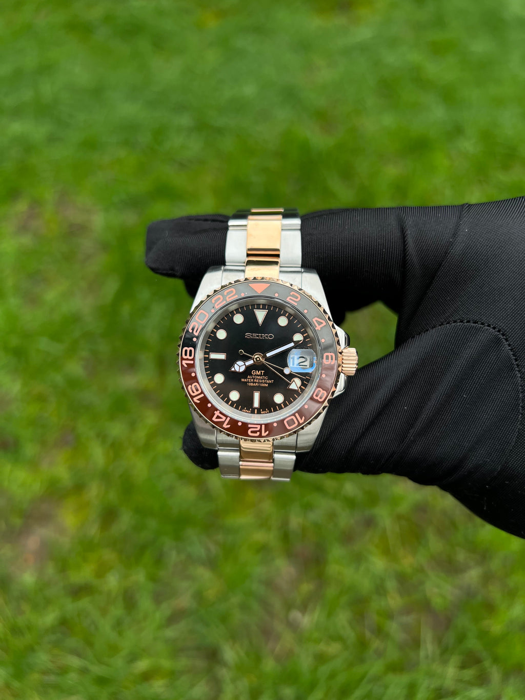 GMT (Rootbeer) Seiko NH34 GMT automatic mod watch. A full watch with a black dial and a black and rose gold bezel on a two tone oyster bracelet held in a glove.