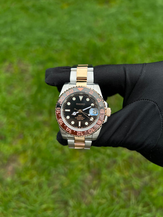 GMT (Rootbeer) Seiko NH34 GMT automatic mod watch. A full watch with a black dial and a black and rose gold bezel on a two tone oyster bracelet held in a glove.