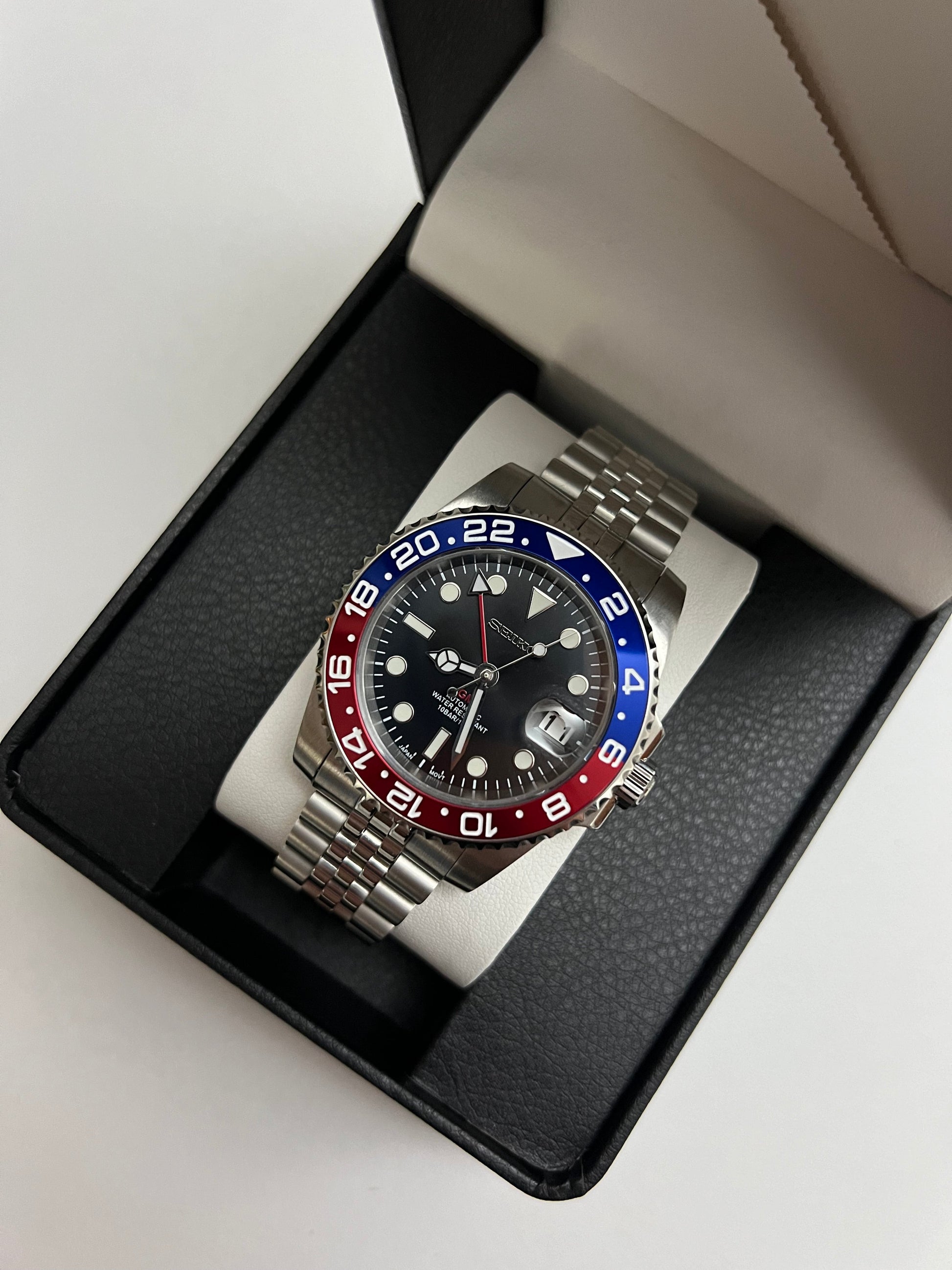 GMT (Pepsi) Seiko NH34 GMT automatic mod watch. A full watch shot inside a box shows a black dial with a blue and red bezel on a silver jubilee bracelet.