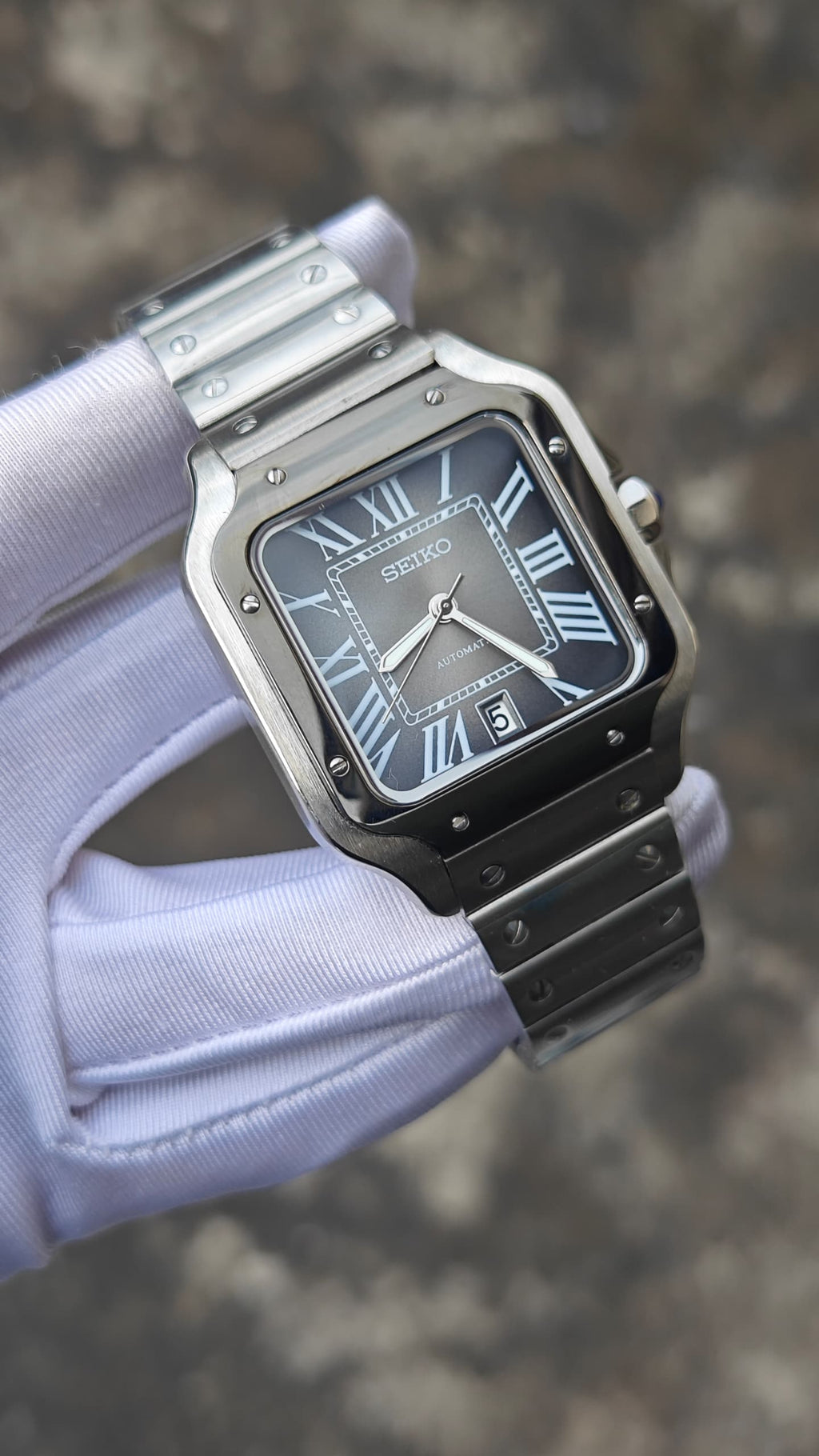 Santos (Graphite) Seiko NH35 automatic mod watch. A full watch shot held by a gloved hand showing a grey dial with a black bezel and a metal link bracelet.